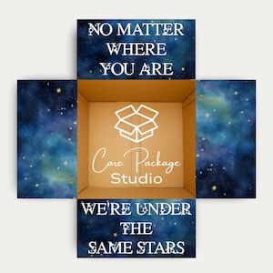 May include: An open cardboard box with a celestial design on the outside. The box is decorated with a starry night sky pattern in shades of blue and gold. Text on the box reads "NO MATTER WHERE YOU ARE" and "WE'RE UNDER THE SAME STARS". The logo for "Care Package Studio" is also visible.
