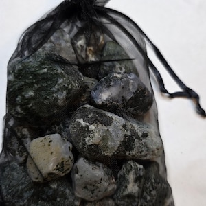 Può includere: Un sacchetto nero a rete con coulisse pieno di pietre verdi e grigie muschiate di varie dimensioni. Le pietre hanno un aspetto strutturato e forme diverse. La borsa è legata in alto con un cordoncino nero.