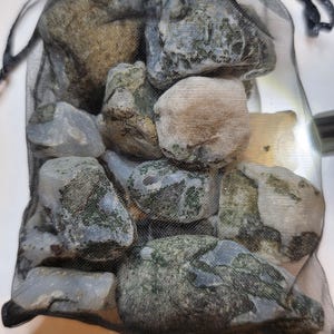 Può includere: Un sacchetto di rete nero pieno di pietre di varie dimensioni. Le pietre hanno un mix di colori, tra cui grigio, marrone e verde. La borsa è chiusa con un cordoncino nero. Le pietre sembrano naturali e hanno una consistenza ruvida.