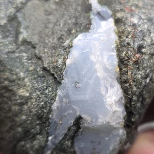 May include: Close-up of a rough, gray rock with a prominent vein of pale blue-gray mineral running through it. The rock's surface is textured, with visible variations in color and pattern.