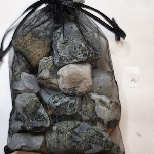 Può includere: Un sacchetto nero a rete con coulisse pieno di varie pietre grigie e verdi. Le pietre hanno un aspetto naturale e strutturato. La borsa è legata in alto con un cordoncino nero. È visibile una piccola etichetta bianca con la lettera "A".