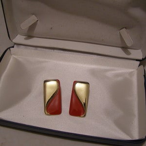 May include: A pair of gold and red earrings in a blue velvet jewelry box. The earrings are rectangular with a curved gold design and a red center.