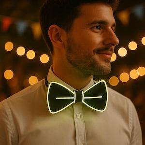 Op de afbeelding: Een zwarte vlinderdas met een heldere neongele omtrek en horizontale middellijnen. De vlinderdas wordt gedragen over een wit overhemd met kraag. De achtergrond is wazig met warme, onscherpe lichten.