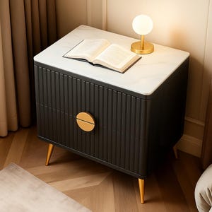 May include: A contemporary black bedside table with a white marble effect top. The table has a ribbed front, gold-coloured drawer handles, and gold-coloured legs. An open book and a small gold lamp with a white globe are on top.