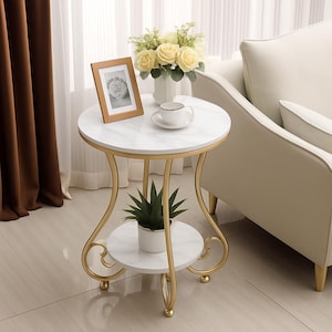 May include: A small side table with a white marble top and a gold-coloured metal frame. The table has a lower shelf and curved legs. A small potted plant, a framed photo, a cup and saucer, and a vase of yellow roses are on the table.