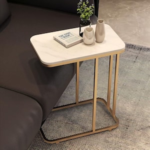 May include: A rectangular side table with a white marble-effect top and gold-coloured metal frame. The table is next to a dark grey sofa and has a book, two vases, and a small plant on top. The table is designed to slide under a sofa.