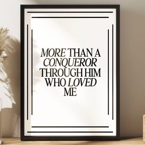 May include: Black framed art print with the text "MORE THAN A CONQUEROR THROUGH HIM WHO LOVED ME" in a bold, elegant font. The print has a white background and is surrounded by a black border with decorative lines. A vase with dried plants is visible on the left.