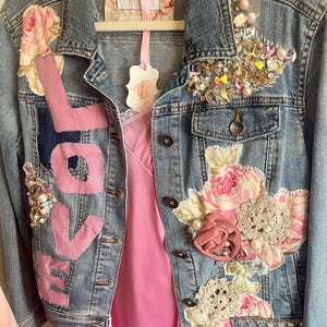 May include: A blue denim jacket featuring pink fabric letters spelling LOVE, floral fabric patches, and intricate embellishments of clear and iridescent crystals, beads, and lace. The jacket has metal buttons and a label reading Farm Charm.