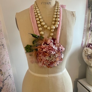 Può includere: Una collana in stile vintage esposta su un manichino. La collana presenta due fili di grandi perle color crema, un nastro rosa e un grande ornamento floreale rosa con foglie verdi e piume. Il manichino è beige chiaro.