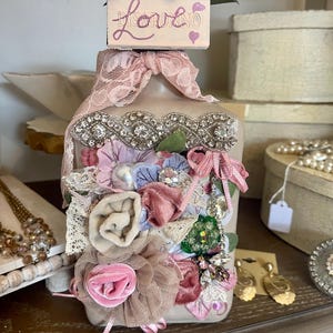 May include: A decorative bottle with a floral and lace design. The bottle is adorned with fabric flowers in shades of pink, beige, and blue, accented with lace and rhinestones. The top features a small box with the word "Love" and two hearts.