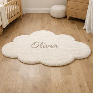 Op de afbeelding: Een crèmekleurig, wolkvormig vloerkleed met de naam "Oliver" geborduurd in een lichtgrijs script. Het vloerkleed ligt op een lichte houten vloer, met een wieg en een dressoir op de achtergrond.