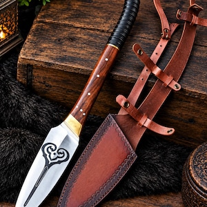 Könnte beinhalten: Ein Dolch mit silberner Klinge, die ein schwarzes herzförmiges Emblem aufweist, einem Holzgriff mit weißen Stiften und einem schwarzen Griffstück. Der Dolch wird von einer braunen Lederscheide mit verzierten Rändern und verstellbaren Riemen begleitet.