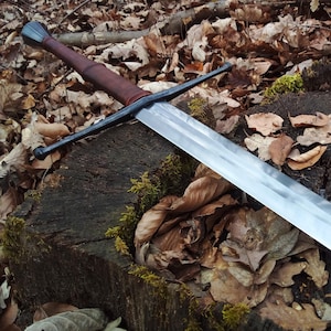 May include: A long, silver sword with a dark brown leather-wrapped handle and a black crossguard and pommel. The sword rests on a weathered tree stump covered in fallen leaves and moss, creating a rustic, outdoor scene.