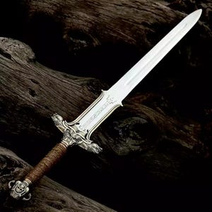 May include: A silver-colored sword with a long, double-edged blade. The hilt features intricate detailing, including animal-like figures and a wrapped brown grip. The pommel has a skull design. The sword is displayed against a dark, textured background.