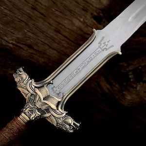 May include: Close-up of a decorative sword with a silver blade and ornate gold-colored hilt. The hilt features detailed carvings and a wrapped handle. The blade has etched text.