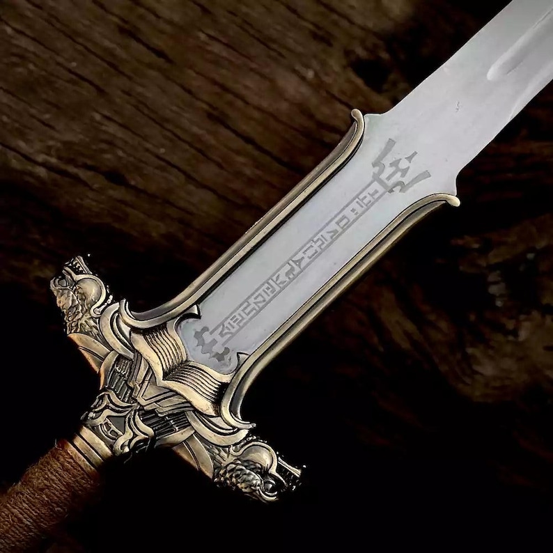May include: Close-up of a decorative sword with a silver blade and ornate gold-colored hilt. The hilt features detailed carvings and a wrapped handle. The blade has etched text.
