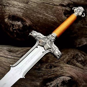 May include: A detailed, silver-colored fantasy sword with an orange wrapped handle. The hilt features dragon and skull designs. The blade has etched text. The sword is displayed against a dark, textured wooden background.