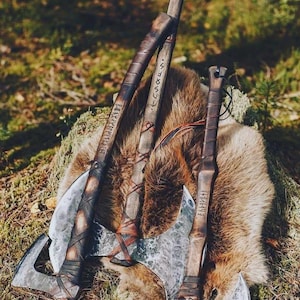 Può includere: Tre asce forgiate a mano con manici in legno scuro avvolti in strisce di cuoio. Le teste in metallo presentano una consistenza martellata e i manici in legno sono incisi con iscrizioni runiche. Le asce sono disposte insieme in un gruppo.