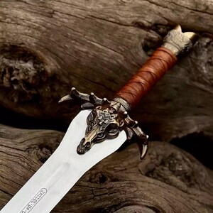 May include: A close-up of a decorative sword with a silver blade and a brown leather-wrapped handle. The hilt features a detailed bronze-colored bull skull design. The sword's pommel has a decorative gold-colored design.