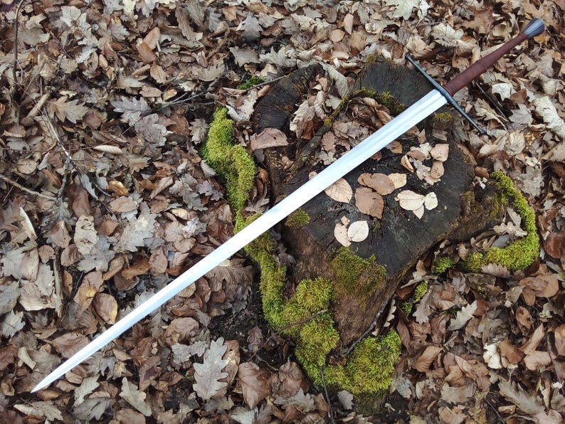 May include: A long, silver sword with a brown handle and crossguard rests on a moss-covered tree stump surrounded by fallen leaves. The sword's blade is straight and sharp, reflecting the light. The overall aesthetic suggests a medieval or fantasy theme.