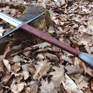 May include: A longsword with a polished steel blade and a brown leather-wrapped handle. The crossguard and pommel are dark metal. The sword rests on a tree stump surrounded by fallen leaves, showcasing a medieval or fantasy theme.