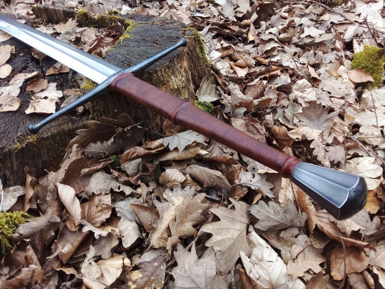 May include: A longsword with a polished steel blade and a brown leather-wrapped handle. The crossguard and pommel are dark metal. The sword rests on a tree stump surrounded by fallen leaves, showcasing a medieval or fantasy theme.
