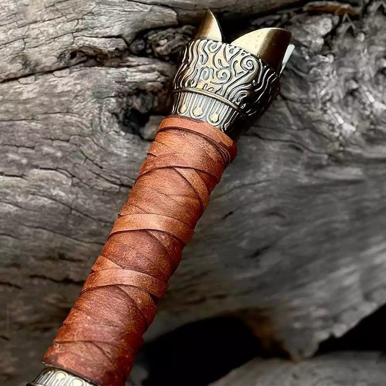 May include: Close-up of a handcrafted axe head with a decorative brass top and a wrapped leather handle. The brass top features an ornate design. The handle is wrapped in brown leather. The axe is likely for decorative or collectible purposes.