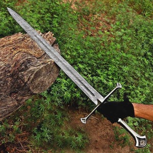 Op de afbeelding: Een lang, zilveren zwaard met een patroon op het lemmet en een gedetailleerde greep. Het zwaard wordt vastgehouden door een gehandschoende hand, met een houten stam en groene bladeren op de achtergrond. De kling is ongeveer 91 cm lang.