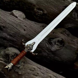 May include: A decorative sword with a silver blade and ornate bronze hilt. The handle is wrapped in brown leather. The sword is displayed against a backdrop of dark, weathered wood.