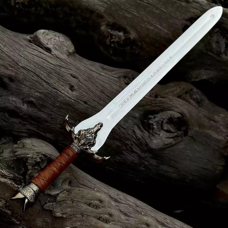 May include: A decorative sword with a silver blade and ornate bronze hilt. The handle is wrapped in brown leather. The sword is displayed against a backdrop of dark, weathered wood.