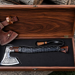 Puede incluir: Un hacha de metal con hoja grabada y mango envuelto en cuero negro, con el nombre Alfzam. Se presenta en una caja de madera rectangular con forro de espuma negra, acompañada de una funda de cuero marrón con broches de presión metálicos.