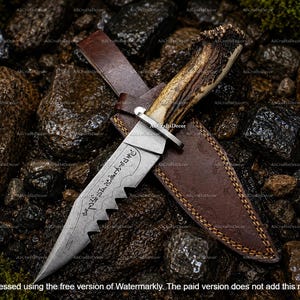 May include: A fixed-blade knife with a textured antler handle and a silver-colored metal guard. The steel blade features a serrated spine and etched runic-style characters. It is accompanied by a brown leather sheath with visible stitching.