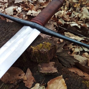 May include: A long, medieval-style sword with a polished silver blade and a dark, textured handle. The sword's crossguard and pommel are black. The sword rests on a weathered wooden surface covered in fallen leaves, creating a rustic aesthetic.