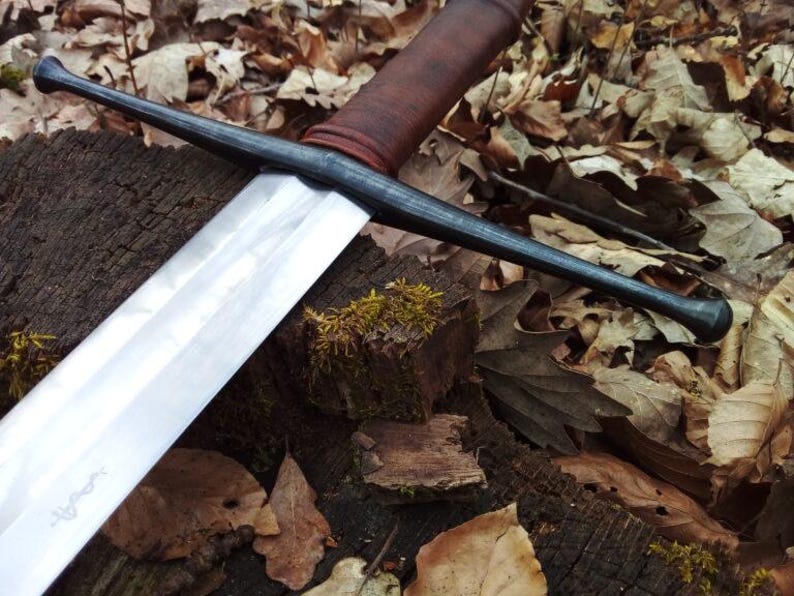 May include: A long, medieval-style sword with a polished silver blade and a dark, textured handle. The sword's crossguard and pommel are black. The sword rests on a weathered wooden surface covered in fallen leaves, creating a rustic aesthetic.