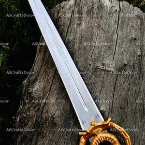 May include: A decorative sword with a silver blade and a gold-coloured handle. The handle features an intricate, circular design. The sword is displayed against a weathered wooden background, with green foliage in the background.