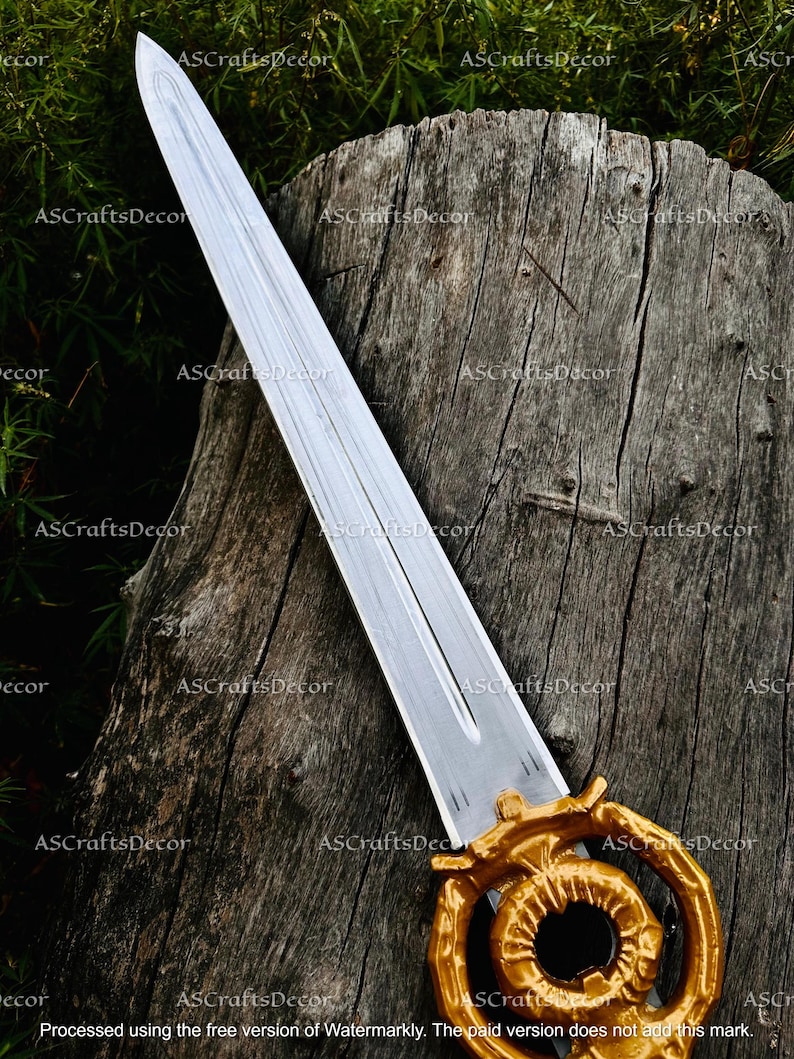 May include: A decorative sword with a silver blade and a gold-coloured handle. The handle features an intricate, circular design. The sword is displayed against a weathered wooden background, with green foliage in the background.