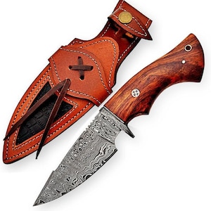 May include: A Damascus steel knife with a wooden handle and a brown leather sheath. The knife blade has a unique wave pattern. The sheath is brown with white stitching and a cross design. The knife is a hunting or utility tool.