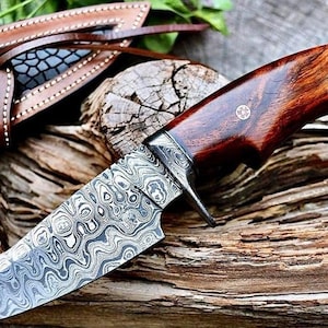 May include: A handcrafted knife with a patterned steel blade and a dark wood handle. The knife rests on a piece of weathered wood, with a leather sheath visible in the background. The blade has a unique, swirling pattern.
