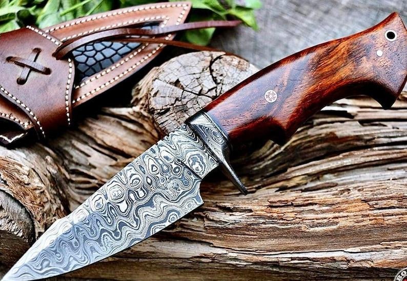 May include: A handcrafted knife with a patterned steel blade and a dark wood handle. The knife rests on a piece of weathered wood, with a leather sheath visible in the background. The blade has a unique, swirling pattern.
