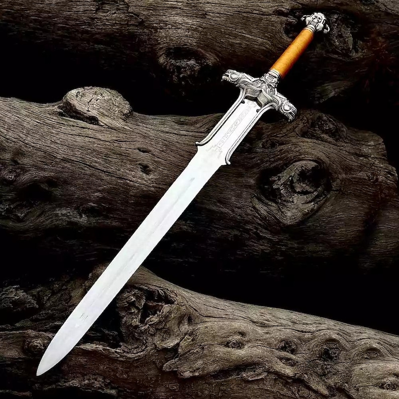 May include: A silver-colored sword with a light brown handle and intricate detailing. The blade is long and sharp, with a polished finish. The hilt features ornate carvings, and the sword rests on a dark wooden surface. A decorative weapon.