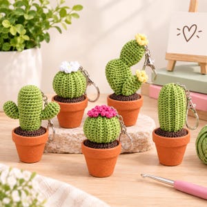May include: Four crocheted cactus keychains in small terracotta-colored pots. Each cactus is green with dark brown soil and features a unique shape, including round, tall, and branched varieties, topped with small white, yellow, or pink flowers.