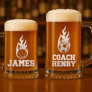 May include: Two clear glass beer mugs filled with beer and foam. One mug has a basketball graphic with the name "JAMES" below it. The other mug has a soccer ball graphic with the name "COACH HENRY" below it. Both mugs have a flame design.