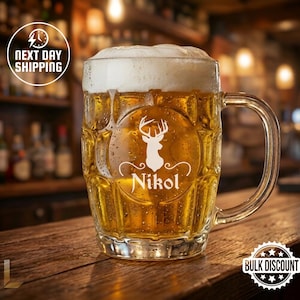 May include: A clear glass beer mug filled with golden beer and a foamy head. The mug features a white deer head design with the name "Nikol" and a handle. The image is set on a wooden bar with a blurred background.