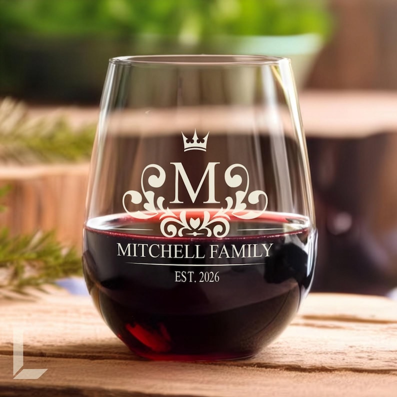 Op de afbeelding: Een helder, steelloos wijnglas gevuld met rode wijn. Het glas heeft een wit ge&euml;tst ontwerp met een kroonpictogram boven de letter M, omringd door krulwerk. Daaronder staat de tekst MITCHELL FAMILY en EST. 2026 in het wit.