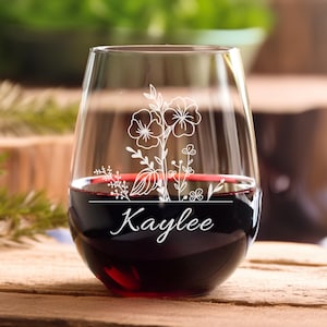 May include: A stemless wine glass filled with red wine. The glass features a white floral design and the name "Kaylee" in cursive. The glass is on a wooden surface.
