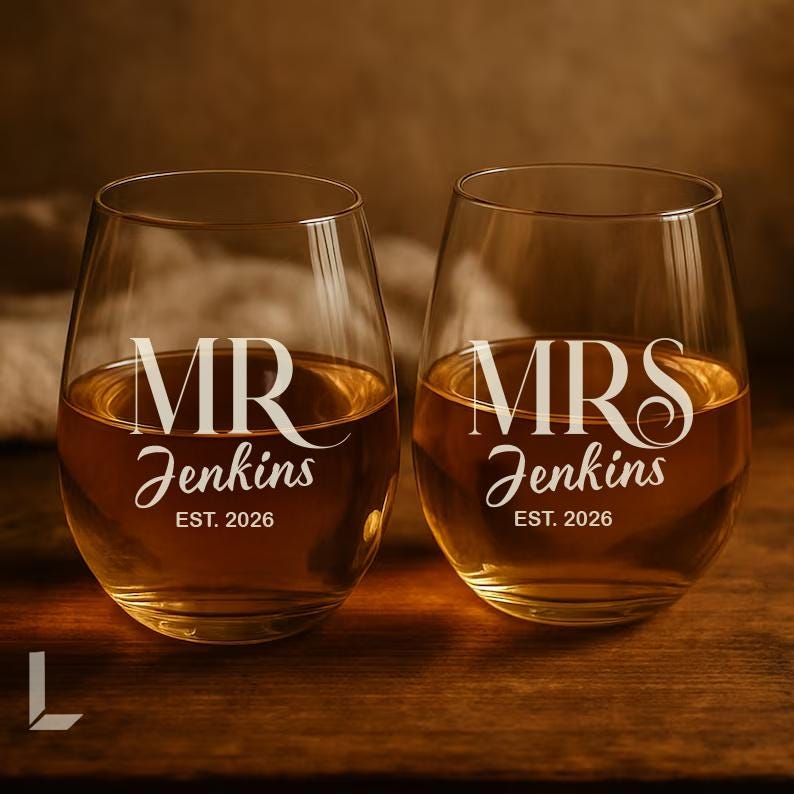 Op de afbeelding: Twee heldere, steelvrije wijnglazen gevuld met een amberkleurige vloeistof. Het linkerglas heeft witte tekst met MR Jenkins EST. 2026. Het rechterglas heeft witte tekst met MRS Jenkins EST. 2026. Beide glazen hebben een ronde, bolle vorm.