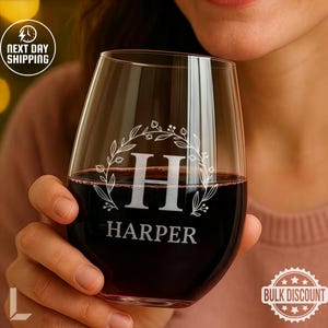 May include: A stemless wine glass filled with red wine. The glass features a white floral wreath design around the letter "H" and the name "HARPER". The glass is held by a person.
