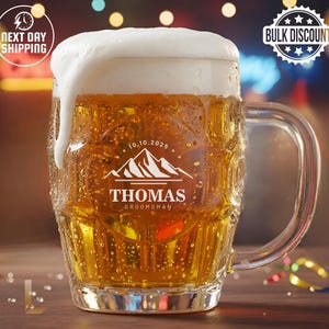 May include: A large, clear glass beer mug filled with golden beer and topped with a thick layer of white foam. The mug has a handle and a personalized design with the name "Thomas" and the word "Groomsman" below a mountain graphic.