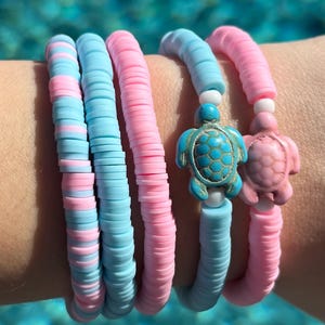 Puede incluir: Cuatro pulseras con temática playera. Dos pulseras son de color azul claro y rosa, y dos tienen dijes de tortuga. Un dije de tortuga es turquesa y el otro es rosa. Las pulseras están hechas de cuentas planas y redondas.