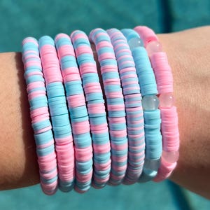 Puede incluir: Una colección de pulseras de colores pastel. Las pulseras están hechas de pequeñas cuentas redondas y planas en bandas alternas de azul claro y rosa. Algunas pulseras incluyen cuentas transparentes y rosas. Las pulseras están apiladas en una muñeca.